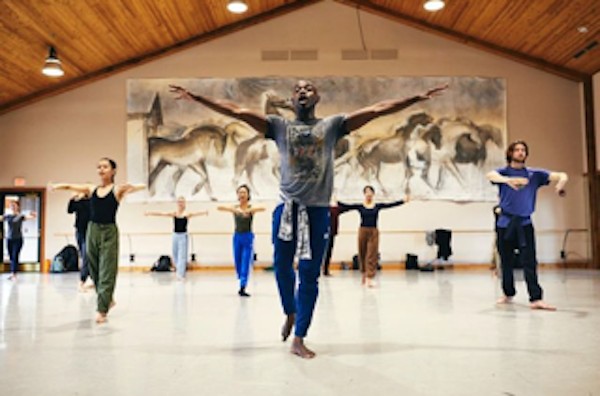 a black man in grey t shirt and navy blue sweats leading a class of 6 people in multi colored practice clothes in a high ceiling studio with a huge mural of horses in black and white behind them